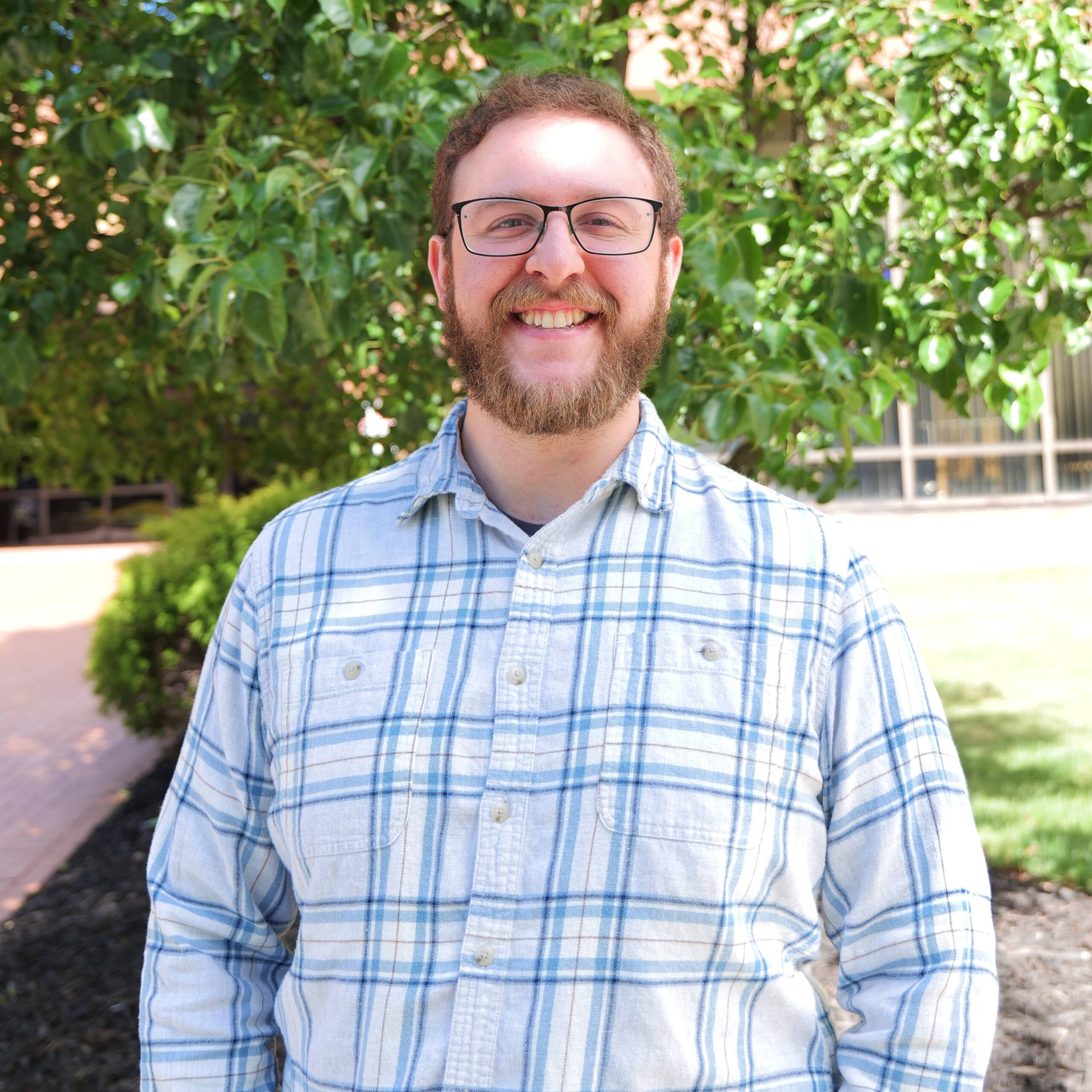 Smiling man with glasses and beard, wearing a blue plaid shirt, outdoors with green bushes behind him, daylight.