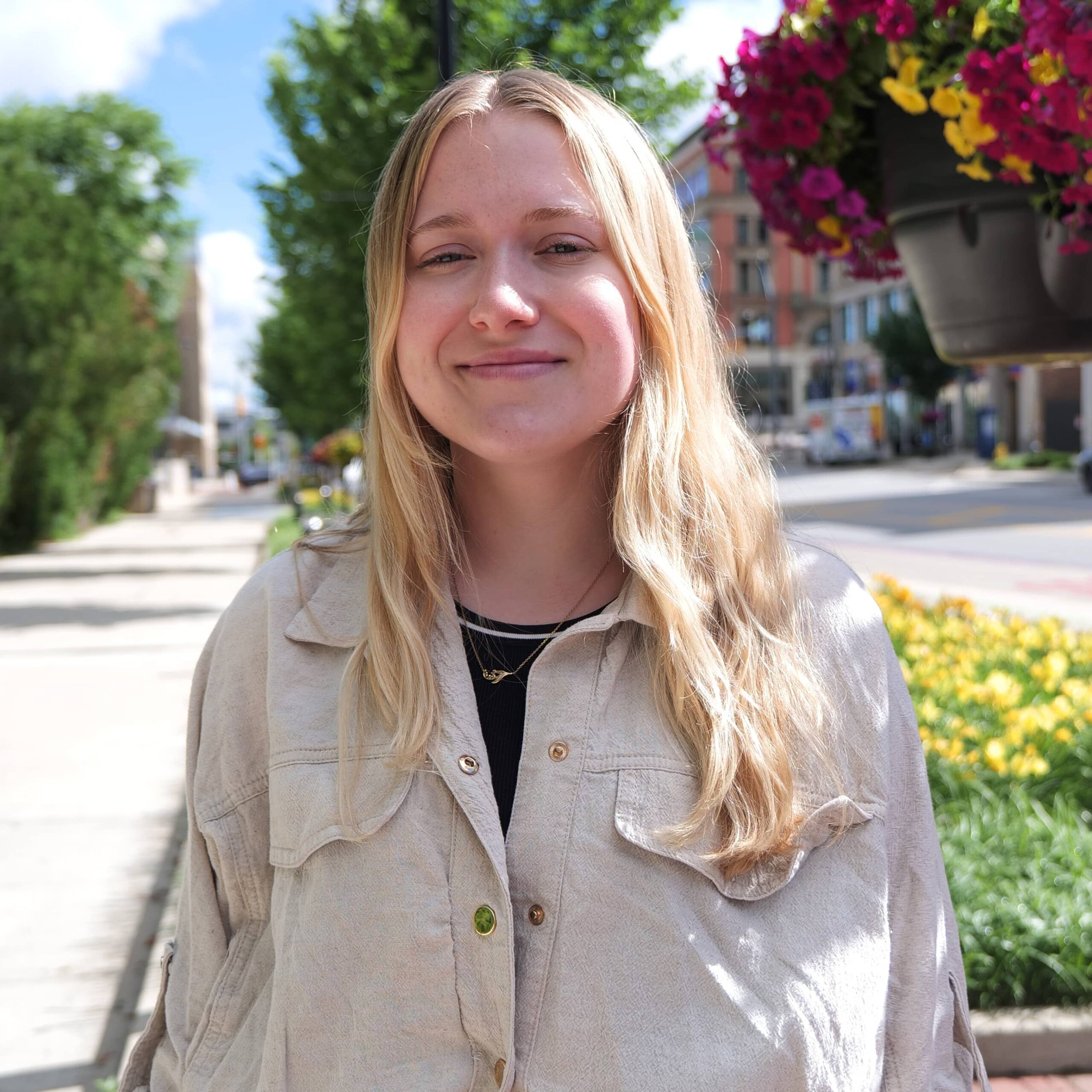 Close-up of a smiling blonde woman wearing a beige jacket outdoors with greenery and hanging flowers in the background.