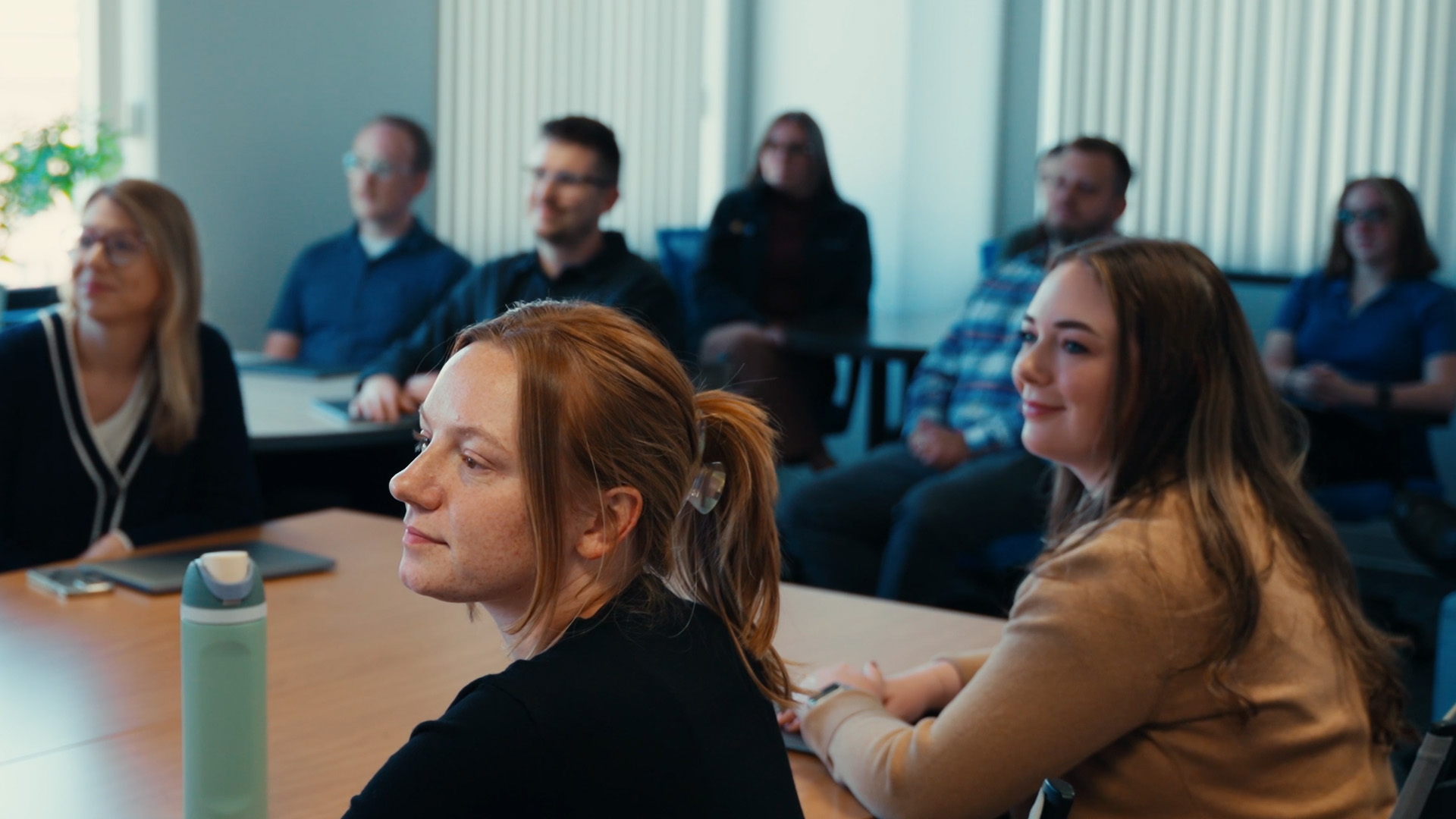 Group of coworkers seated around a conference table, some looking toward the front of the room during a meeting in a bright office.