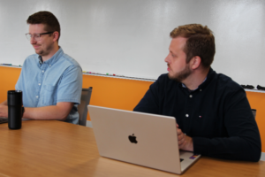 Two men sit at a wooden table with laptops; left man in a light blue shirt and glasses, right man in a dark shirt using a MacBook, whiteboard behind them.