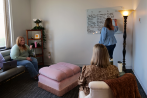 Three women in a small office meeting; one writes on a whiteboard while two talk, pink tufted ottoman in the center.