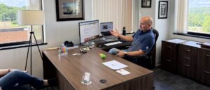 Man in a blue polo sits at a large wooden desk in a bright office, gesturing while talking to someone off-screen, with dual computer monitors in front of him.