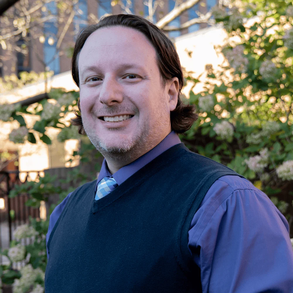Smiling man outdoors, wearing a purple shirt and navy vest, with greenery in the background.