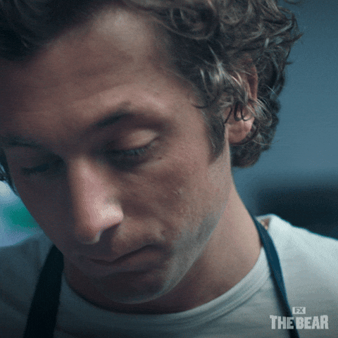 Jeremy Allen White Cooking GIF by The Bear – FSM – Digital Marketing Agency Close-up of a man looking downward with a somber expression, wearing a white shirt and a dark lanyard.