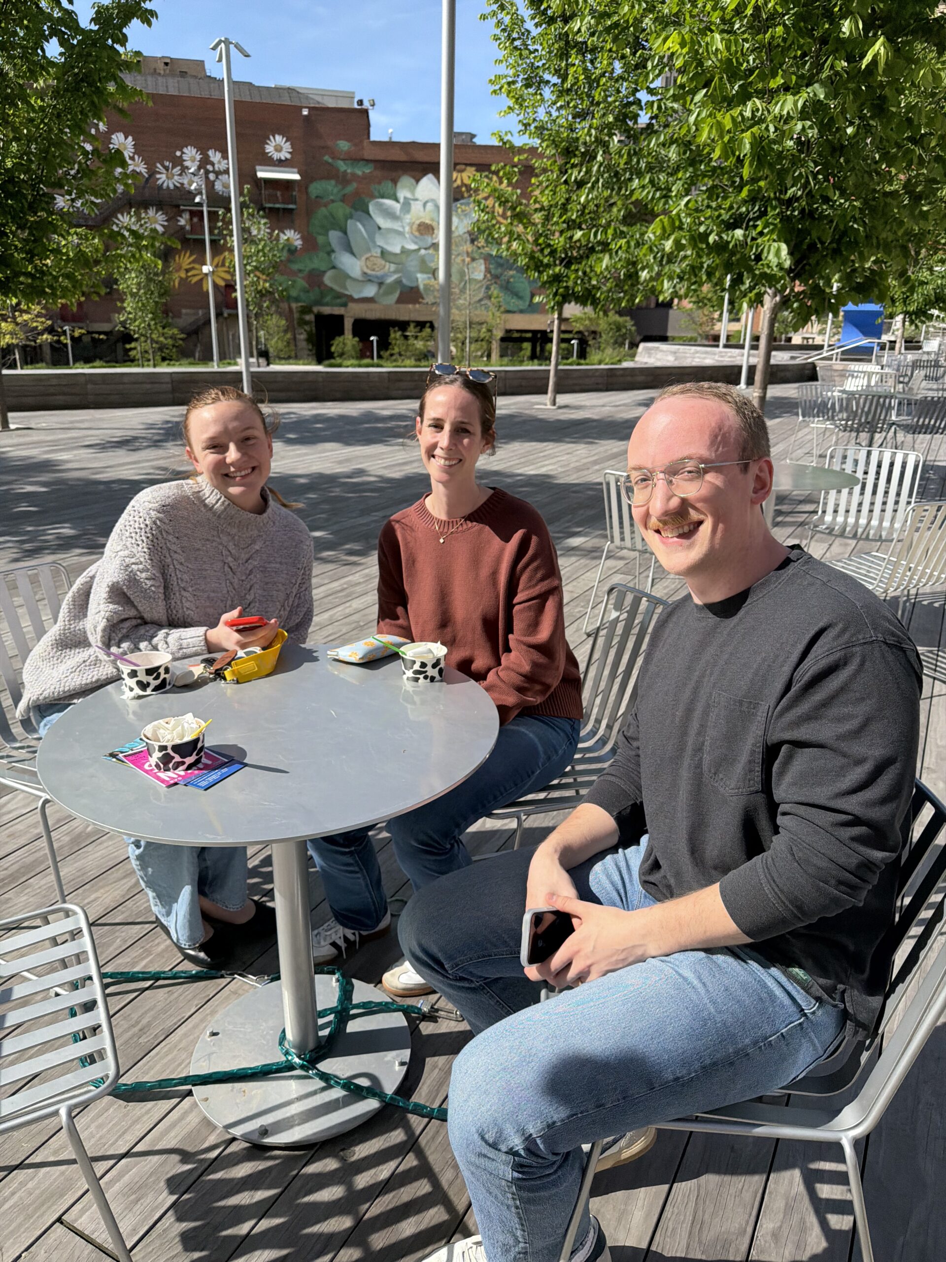 Five women sitting on a wooden bench with drinks in their hands and office buildings behind them.