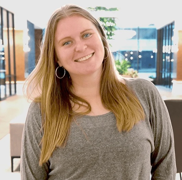 Smiling woman with long blond hair in a gray top standing in a bright indoor lobby.
