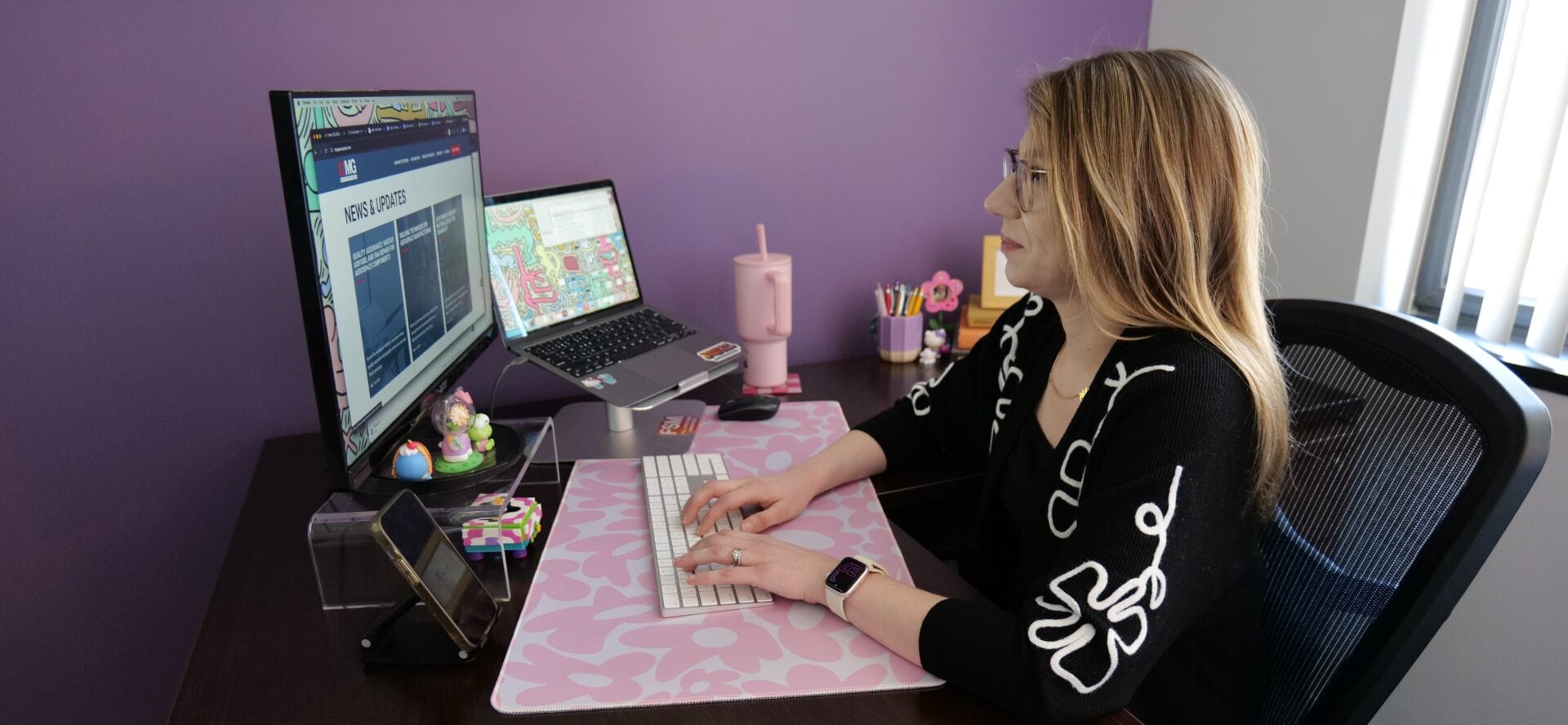 P1011656 Woman working at a desk with dual monitors on a purple wall, typing on a keyboard, pink accents nearby.