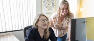 Two coworkers collaborate at a desk, smiling at a computer screen; one seated with glasses, the other standing beside her.