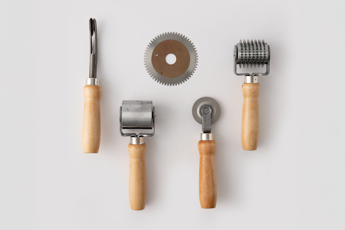 Four industrial hand tools on white: wood-handled seam probe, serrated blade, spiked roller, and metal stitcher.