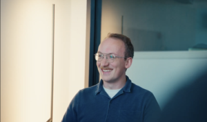 Smiling man with glasses in a blue polo shirt seated in a bright office, engaged in conversation.