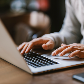 Close-up side view of hands typing on a silver laptop keyboard on a wooden table with a soft, blurred background.