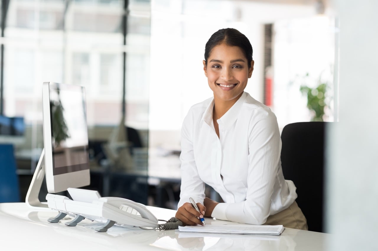 PortraitOfBeautifulYoungBusinesswomanSittingAtHerDeskIn – FSM - Akron Ohio Marketing Agency Woman in a white blouse sits at a bright office desk with a computer and phone, smiling at the camera.